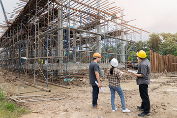 Team of engineers and architects wearing safety helmets discussing building progress at construction site with scaffolding structure.