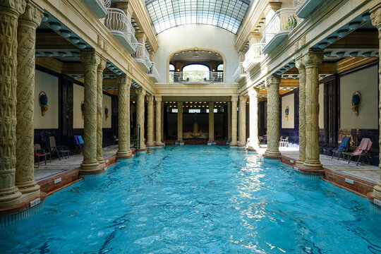 View of the Gell&Atilde;&copy;rt Thermal Bath pool shimmers under a glass ceiling, framed by ornate columns and dark wood accents, Budapest, Hungary.
