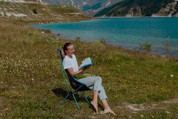 A young Caucasian woman with long brown hair sits on a chair by a lake, reading a book. The background features mountains and clear blue water.
