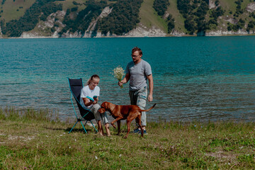 Couple enjoying a sunny day by a lake in the Caucasus. Girl is sitting in a chair, while man stands with a dog and flowers. Green hills are in the background.