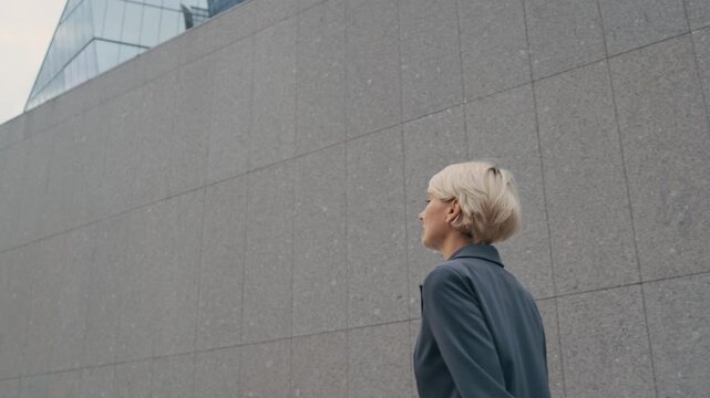 Low angle tracking shot of confident business lady wearing gray blazer walking along outdoor wall in modern city center