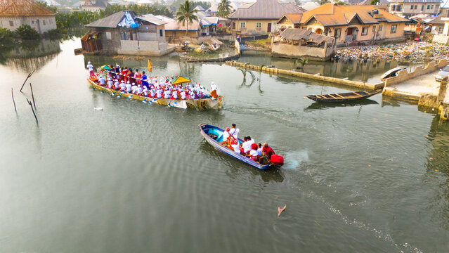 Aerial view of Niger Delta War Canoes gliding through the murky waters, carrying warriors in white robes amidst the stilt houses, Ogu Town, Rivers State, Nigeria.