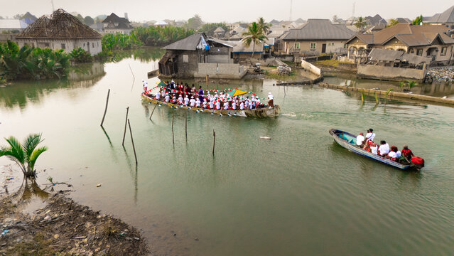 Aerial view of War Canoe of the Niger Delta carrying warriors navigate through waterways surrounded by houses, Ogu Town, Rivers State, Nigeria.