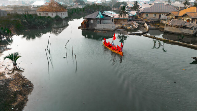 Aerial view of a vibrant red-clad group gliding on a golden boat through the dark waters, surrounded by rustic buildings, Nigeria.