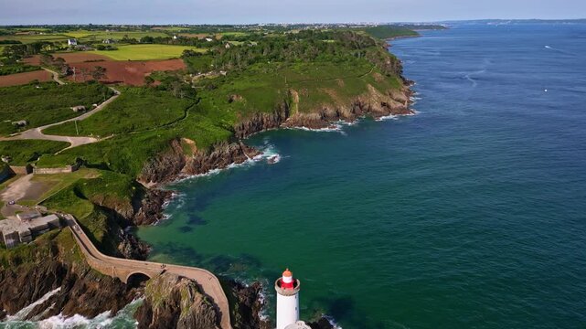Drone pulling away from Phare du Petit Minou lighthouse in Plouzan&eacute;, Brittany, France, showing the white lighthouse on rocky cliffs, turquoise ocean, and lush green landscape under a bright sky.