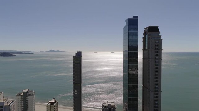 Drone shot flying through and showcasing Balne&aacute;rio Cambori&uacute;&rsquo;s skyline with YachtHouse by Pininfarina, One Tower and Millennium Palace, set against the Atlantic Ocean and distant islands.
