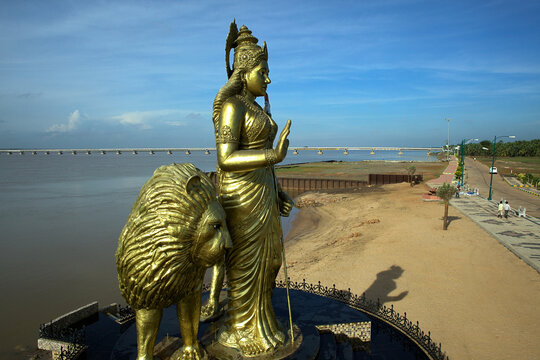 Aerial view of golden sculpture of a woman and a lion, standing proudly against the wide river and distant bridge, shimmering in the sunlight, Vijayawada, Andhra Pradesh, India.