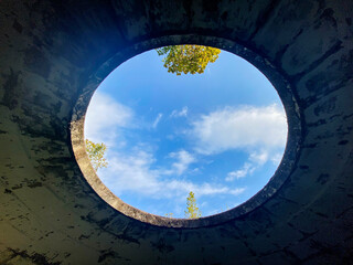 A round hole in a cracked concrete ceiling reveals a view of blue sky with clouds and treetops