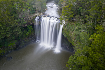 Aerial view of the cascading waterfall plunges into the dark pool below, framed by the lush, vibrant greens of the surrounding forest, Kerikeri, Northland Region, New Zealand.