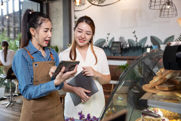 Female barista showing a digital tablet to a customer inside a cozy café, discussing menu options near the pastry counter with fresh bread and cakes displayed, highlighting modern small business