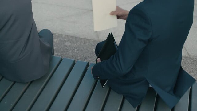Cropped shot of unrecognizable businessman handing document to woman exchanging it to external data drive while sitting on bench during outdoor confidential meeting