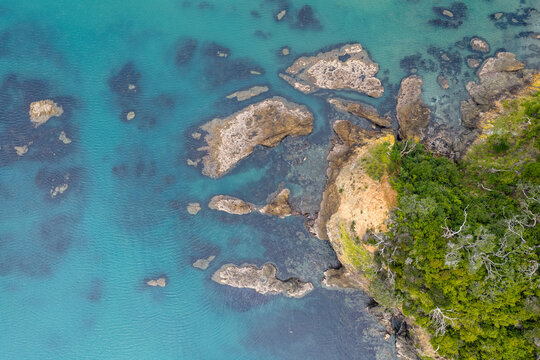 Aerial view of the rugged coastline where turquoise waters meet rocky outcrops and verdant cliffs, Helena Bay, Northland, New Zealand.
