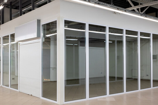 Empty retail space with glass walls and closed white roller shutter inside modern commercial building. 