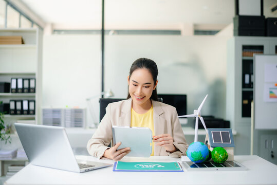 Asian businesswoman in office planning green energy with wind turbine, solar panel, globe. Concept of sustainability, climate change, and reducing CO2 emissions.