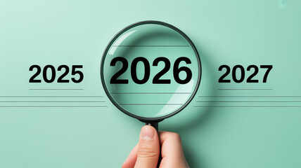 Conceptual image with years 2025, 2026, 2027 in a row on pastel green background. A hand holds a magnifying glass over 2026, enlarging it to symbolize focus, planning, and reflection