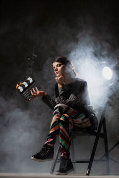 Female movie director on set with smoke background. Girl holding clapperboard on fire.