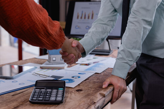 Business partners shaking hands over office desk after signing a contract for a new house purchase, with a miniature house, calculator, and financial documents visible