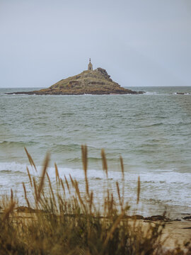 View of a distant, rocky islet crowned with a lighthouse, framed by golden reeds swaying in the foreground breeze, ilot Saint-Michel, Bretagne, France.