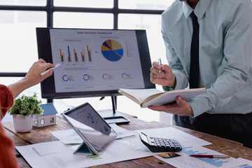 Business colleagues analyzing sales charts and graphs displayed on a computer screen and tablet during a collaborative working session, taking notes and discussing data