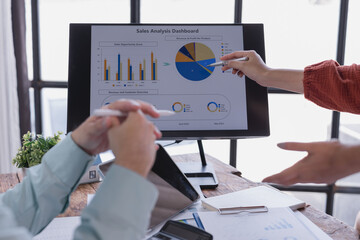 Two businesswomen are discussing sales data displayed on a computer screen, pointing at charts and graphs with pens, analyzing revenue and customer overview in a collaborative office environment