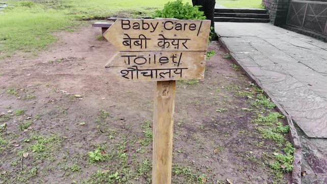 Tiltup shot showing a wooden bilingual direction signpost showing arrows for Baby Care and Toilet facilities, written in both English and Hindi in a public area with greenery and stone pavement.