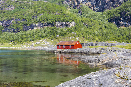 View of a vibrant red cabin reflecting on the still, clear waters, nestled amidst rocky shores and lush green mountains in Mjelde, Bodo, Nordland, Norway.