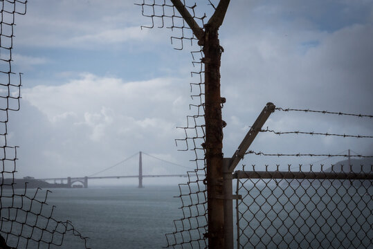 View of the Golden Gate Bridge fading into the misty horizon beyond a weathered, rusted chain-link fence, its wire frayed and broken, San Francisco, California, United States.