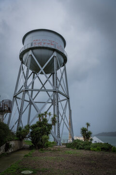 View of a towering water tank against a moody sky, with Alcatraz Island visible in the distance, a stark contrast of industrial and natural beauty, San Francisco, California, United States.