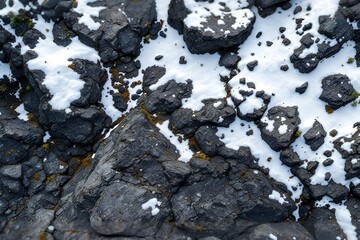 Textured dark volcanic rocks partially covered with patches of white snow, showcasing a stark and rugged natural landscape in a close-up, high-angle view