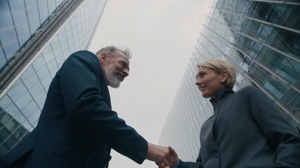 Low angle view of adult Caucasian woman shaking hands with mature male business partner outside glass office buildings in city - Powered by Adobe