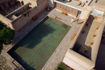 Nagaur, India - 19 October 2010: Aerial view of a serene rectangular swimming pool reflecting the sky, nestled within the peach-toned walls of a traditional Rajasthani palace.