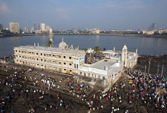 Mumbai, India - 25 October 2006: Aerial view of the Haji Ali Dargah, a striking white structure amidst the dark rocks, surrounded by a sea of devotees under a hazy sky.