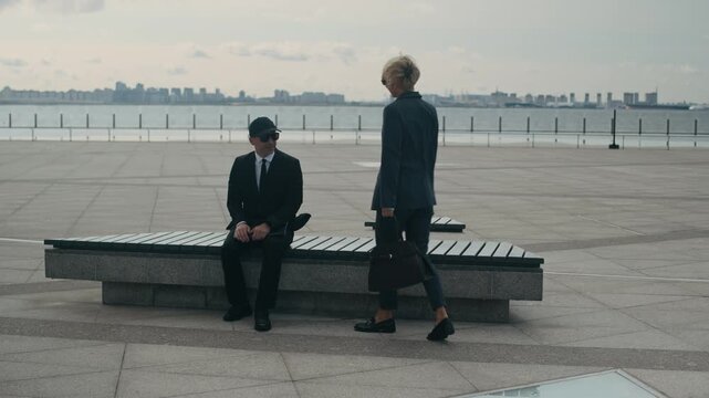 Wide stab shot of adult business lady in suit exchanging secret documents with man in cap and sunglasses sitting on bench near waterfront during outdoor confidential meeting