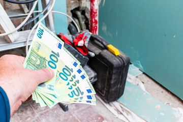 Expensive renovation of an old apartment, a man's hand holding a bundle of 100 euro paper money, construction tools in the background.