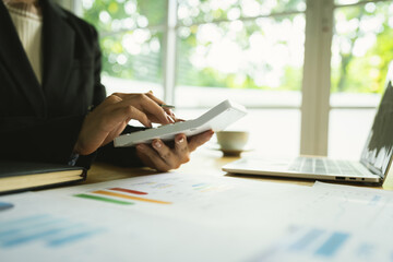 A businessman calculates finances using a calculator, analyzing charts and graphs on documents, while working with laptop and smartphone in office.