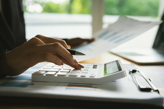 A businessman calculates finances using a calculator, analyzing charts and graphs on documents, while working with laptop and smartphone in office. - Powered by Adobe