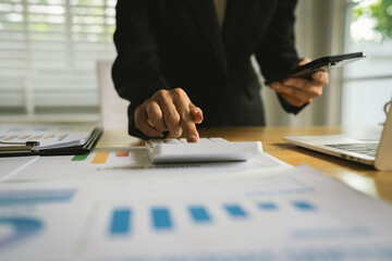 A businessman calculates finances using a calculator, analyzing charts and graphs on documents, while working with laptop and smartphone in office.