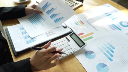 A businessman calculates finances using a calculator, analyzing charts and graphs on documents, while working with laptop and smartphone in office. - Powered by Adobe