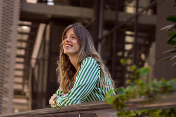 Smiling Woman Leaning on Railing in Green Striped Shirt Outdoors. Portrait of a confident young businesswoman in front of the office building