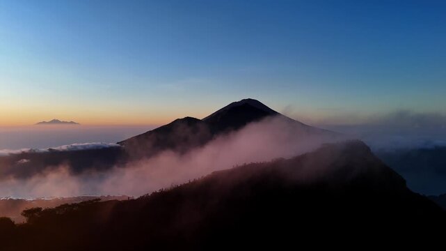 A golden sunrise cast light over Mount Batur in Bali, Indonesia, with hikers gathered at the summit to witness the mist veiling volcanic peak and glowing clouds.