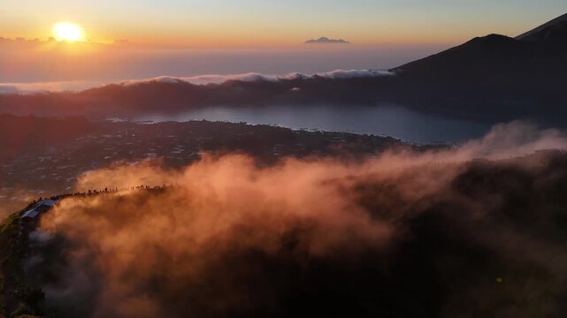 A golden sunrise cast light over Mount Batur in Bali, Indonesia, with hikers gathered at the summit to witness the mist veiling volcanic peak and glowing clouds.