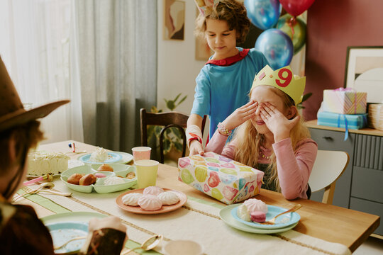Caucasian girl wearing birthday crown with number nine sitting at table, covering eyes while another Caucasian child standing behind presenting wrapped gift during birthday celebration