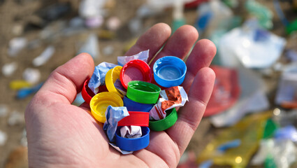 A hand holds a handful of colorful, discarded plastic bottle caps over a messy, littered ground. The image is a powerful symbol of plastic pollution and the importance of recycling plastic waste.