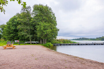 Raindrops ripple across lake beside sandy beach with wooden pier picnic tables grill and green trees. Sweden.