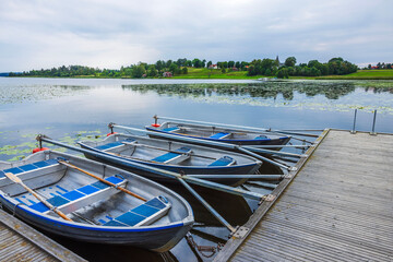 Rowboats tied to wooden pier on calm lake with green shore and village houses visible in...