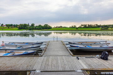 Wooden pier with boats on calm lake against green fields trees and village houses in background....
