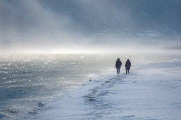 Fototapeta premium Winter walk along the snowy beach with dramatic light and two figures silhouetted against the misty horizon.