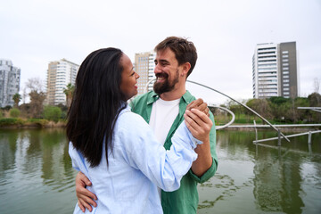 Interracial couple holding hands and dancing together near lake in urban park, enjoying romantic moment smiling. Image with copy space.
