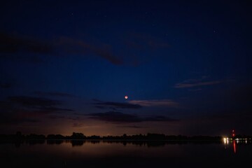 Blood moon shines over calm water reflecting distant lights under a dark blue night sky with clouds