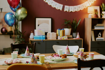 Birthday party table set with colorful plates, cups, party hats, sweets and pastries in decorated living room, balloons and wrapped gifts arranged on cabinet in background